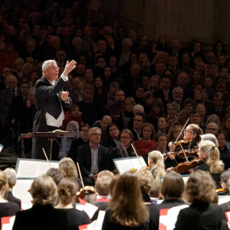 Andrew Carwood, Director of Music, conducts an orchestra with a packed audience in the background. Andrew Carwood, Director of Music, conducts an orchestra with a packed audience in the background.