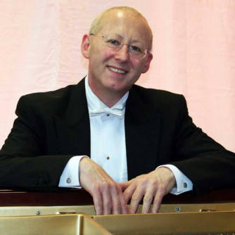 Nicholas Walker, wearing a black suits, leans on a piano smiling