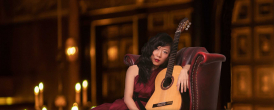 Chinese classical guitarist Xuefei Yang sits next to her guitar on a plush maroon leather armchair. In the background there are lit candelabra in the Sam Wanamaker Playhouse.